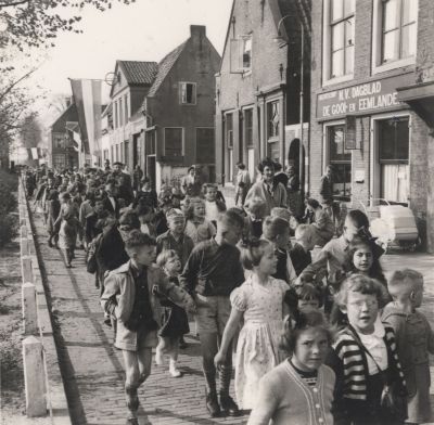Optocht-van-schoolkinderen
Optocht van schoolkinderen.
Rechts kantoorboekhandel van Rijswijk-den Dekker en het postkantoor.
Tevens agentschap van de Gooi-en Eemlander.
In het pand met de vlag de winkel/werkplaats van Schilder Hennipman.
Trefwoorden: Centrum Middenstand.