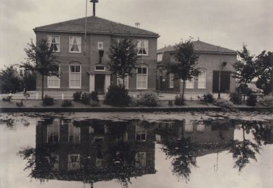 Gemeentehuis-NdB
Gemeentehuis voor 1975  demping Reevaart.  Op het dak van het gemeentehuis de  brandweer  sirene. Naast het gemeentehuis het politiebureau en de brandweerkazerne. Achter de kazerne de slangenmast waarin de brandslangen gehesen werden om te drogen na gebruik. In 1960 sloeg een slang los door de storm en vernielde een raam van de woning boven de kazerne. Zie ook foto 039 s 5-12-1, 1A, 2 en 2A
