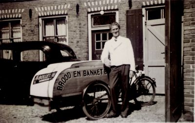 Bakker-Arie-Roukens-met-zijn-bakfiets
Bakker Arie Roukens bij de bakfiets aan de achterzijde van het pand Overmeerseweg 110.
Links woonhuisramen en de luiken van de bakkerij.
Met deze bakfiets ging hij met brood en banket langs zijn klanten.
Trefwoorden: Bakkerij